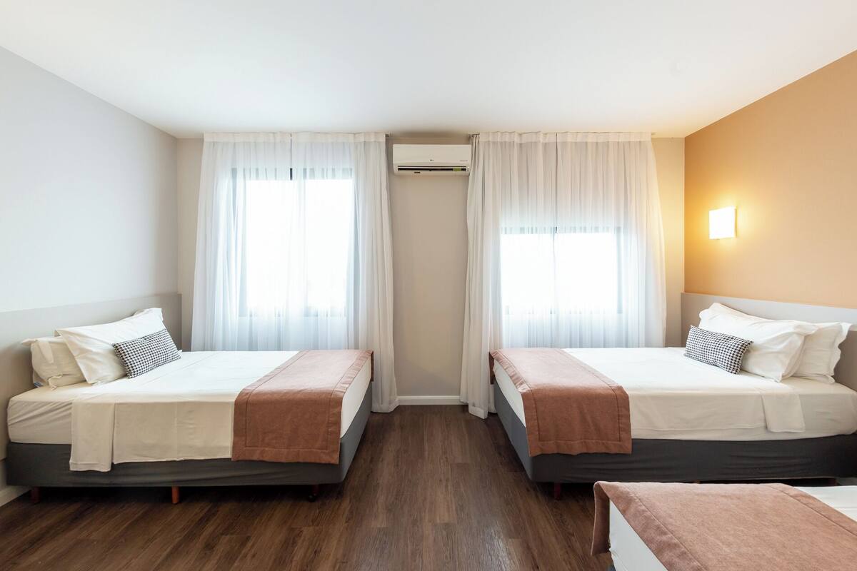 A hotel room with two beds, a wooden floor, a wall-mounted air conditioner, and a light fixture.