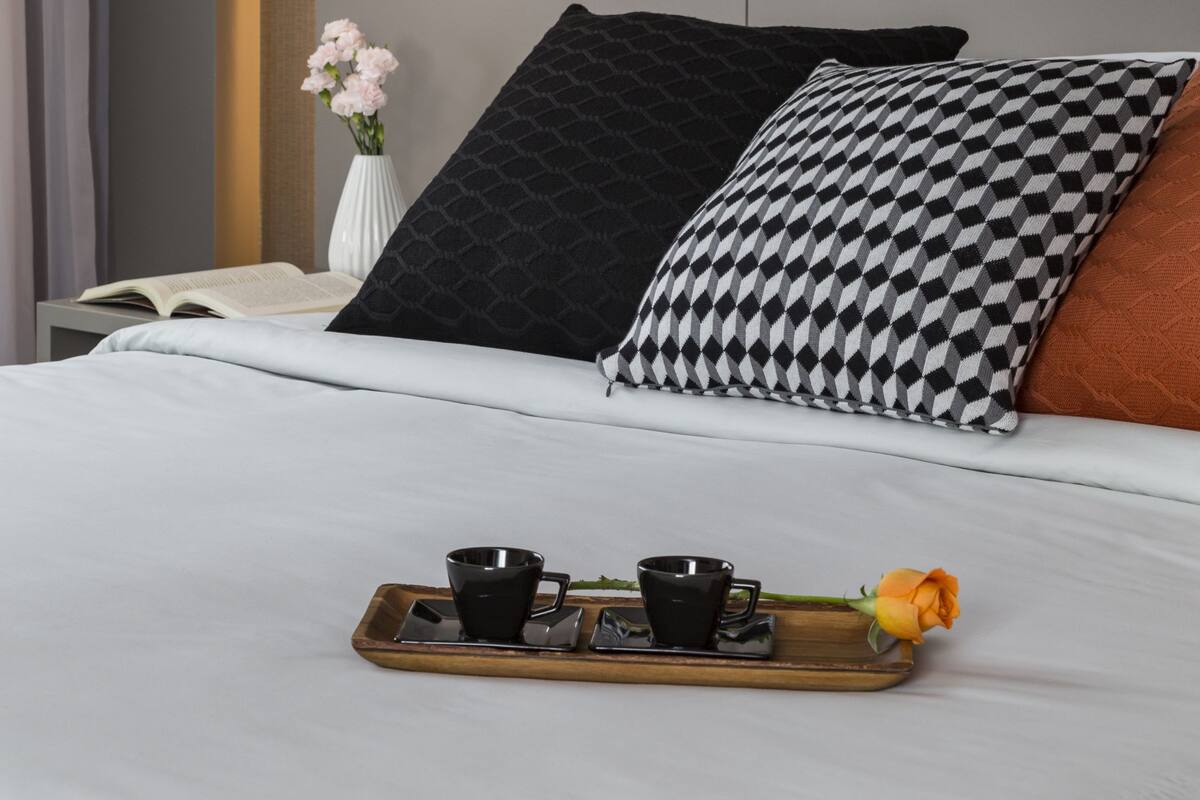 Un lit bien fait, avec un plateau contenant des tasses de café et une rose, une lampe de chevet et un vase rempli de fleurs.