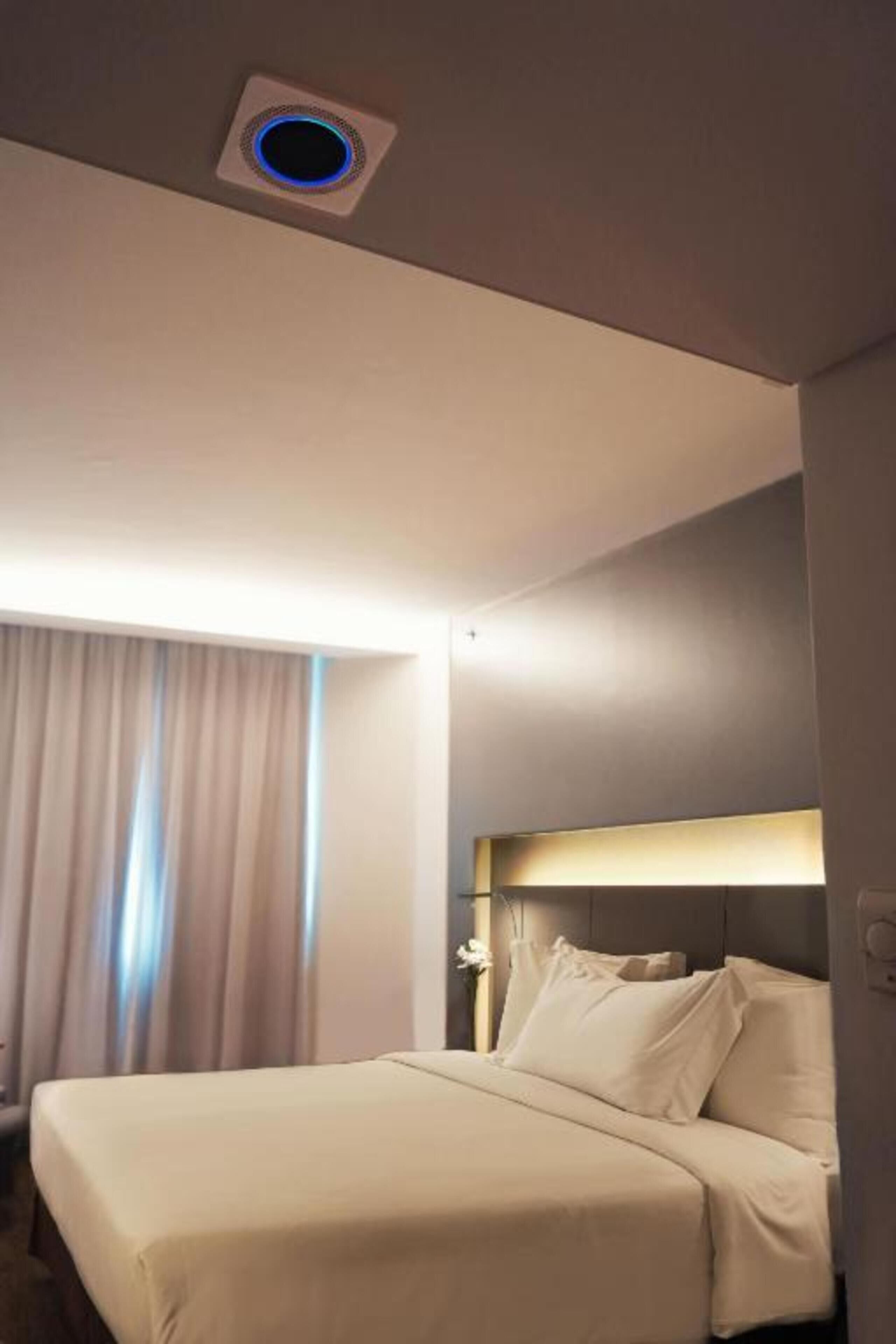 A hotel room with a bed, a headboard, a window with curtains, and a ceiling light.