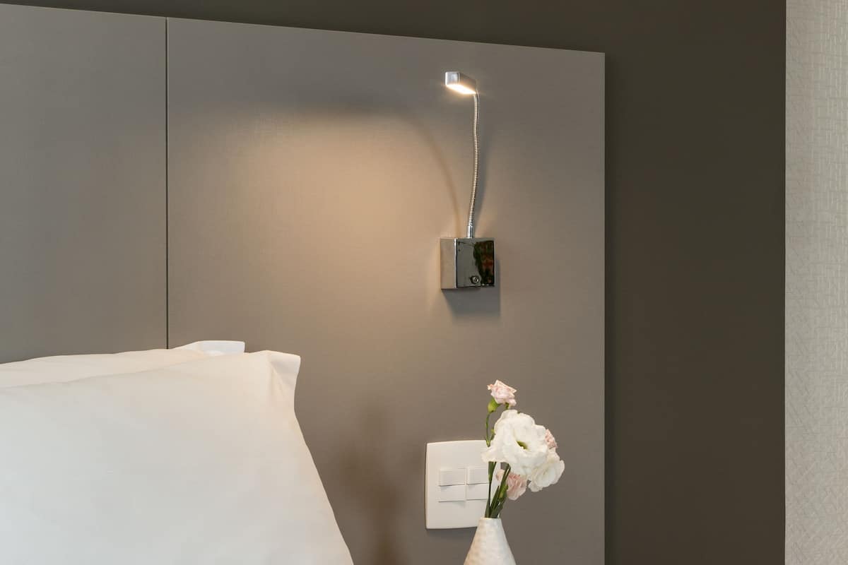 Une chambre d’hôtel moderne, équipée d’un lit, d’une table de chevet ornée d’un vase de fleurs et d’un luminaire fixé au mur.