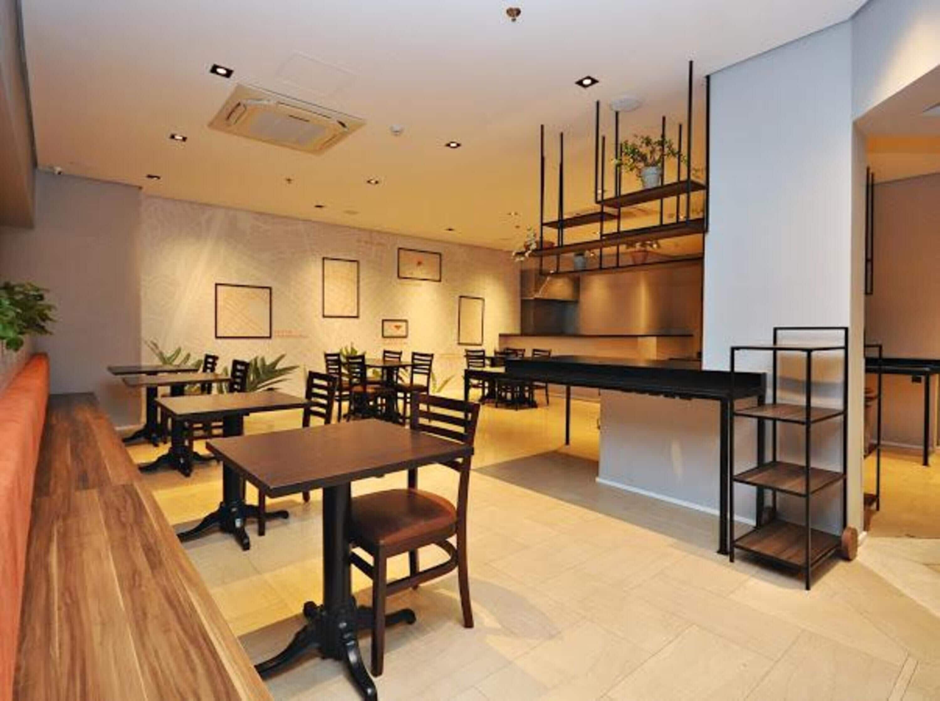 Un área de comedor moderna con mesas y sillas de madera, una cocina al fondo y una escalera que sube a un nivel superior.