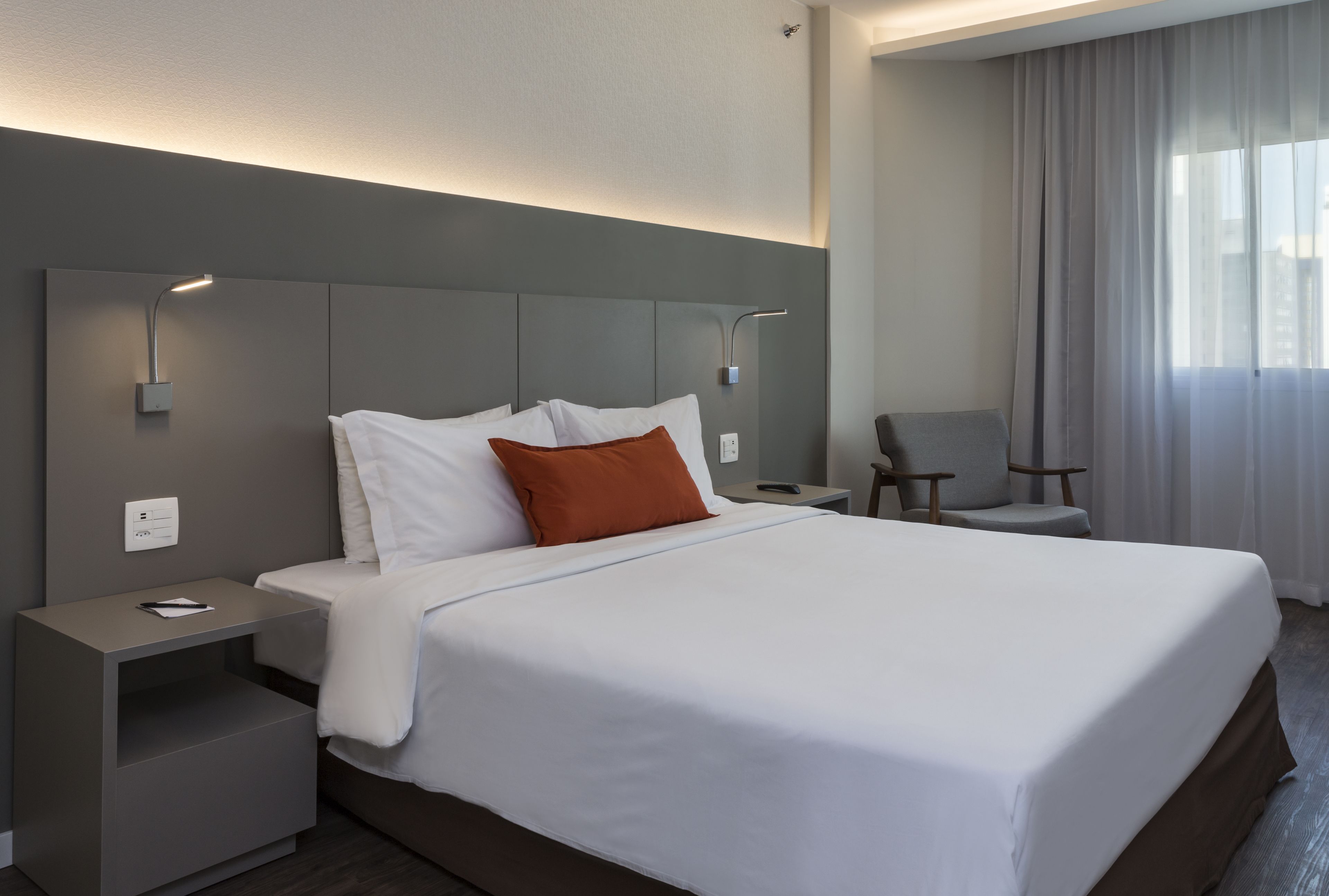 A modern hotel room with a large bed, a nightstand, a chair, and a window with curtains.