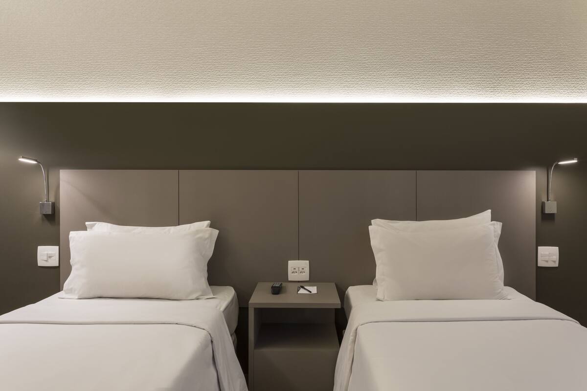 Une chambre d’hôtel avec deux lits simples, une petite table de chevet avec une télécommande et un stylo, et des lampes fixées au mur.