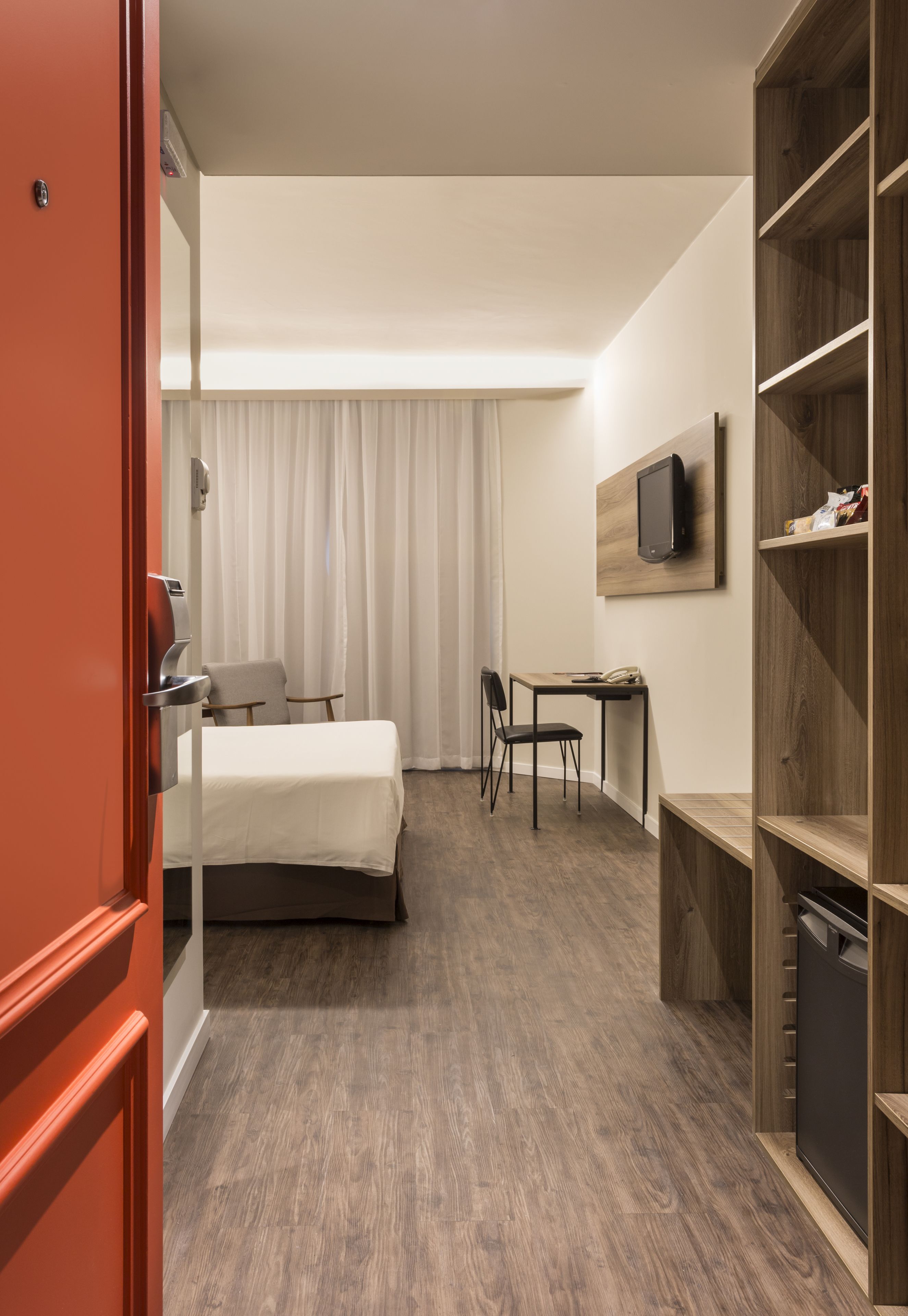 Habitación de hotel con cama, escritorio, silla, televisor montado en la pared y armario.