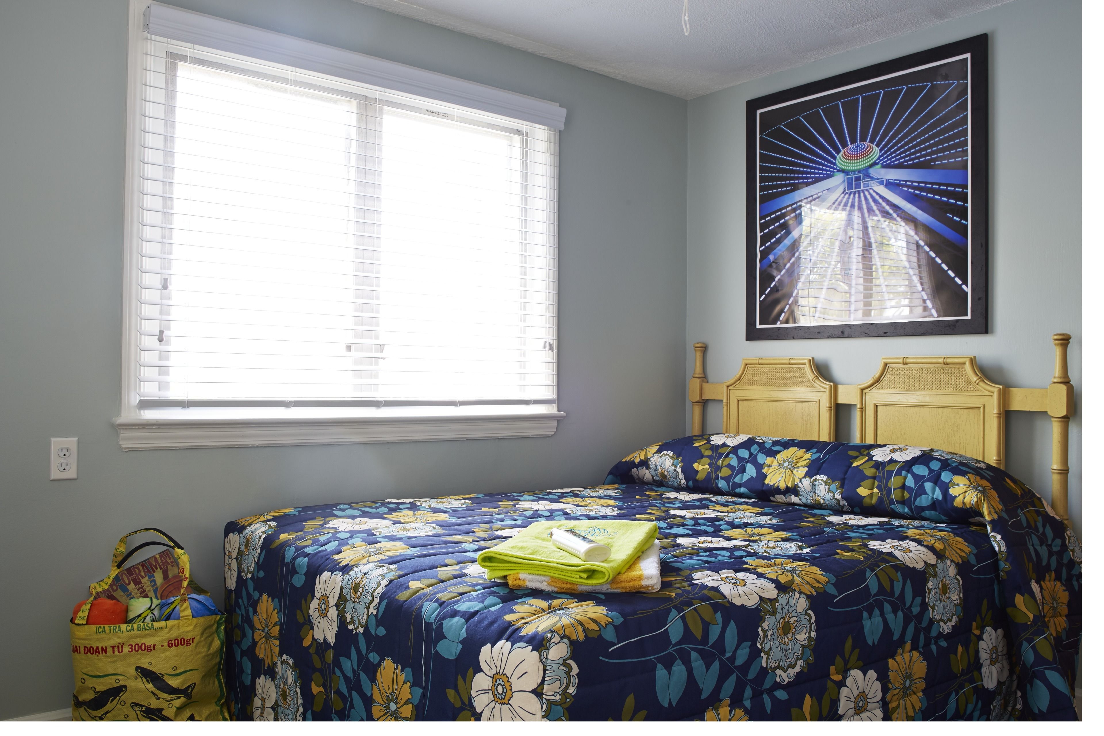 Un dormitorio con un ventanal grande, una cama con ropa de cama floral, un cuadro en la pared y un bolso pequeño en el suelo.