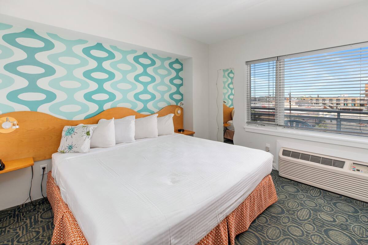 Quarto de hotel com uma cama grande, parede estampada, janela com persianas e radiador.