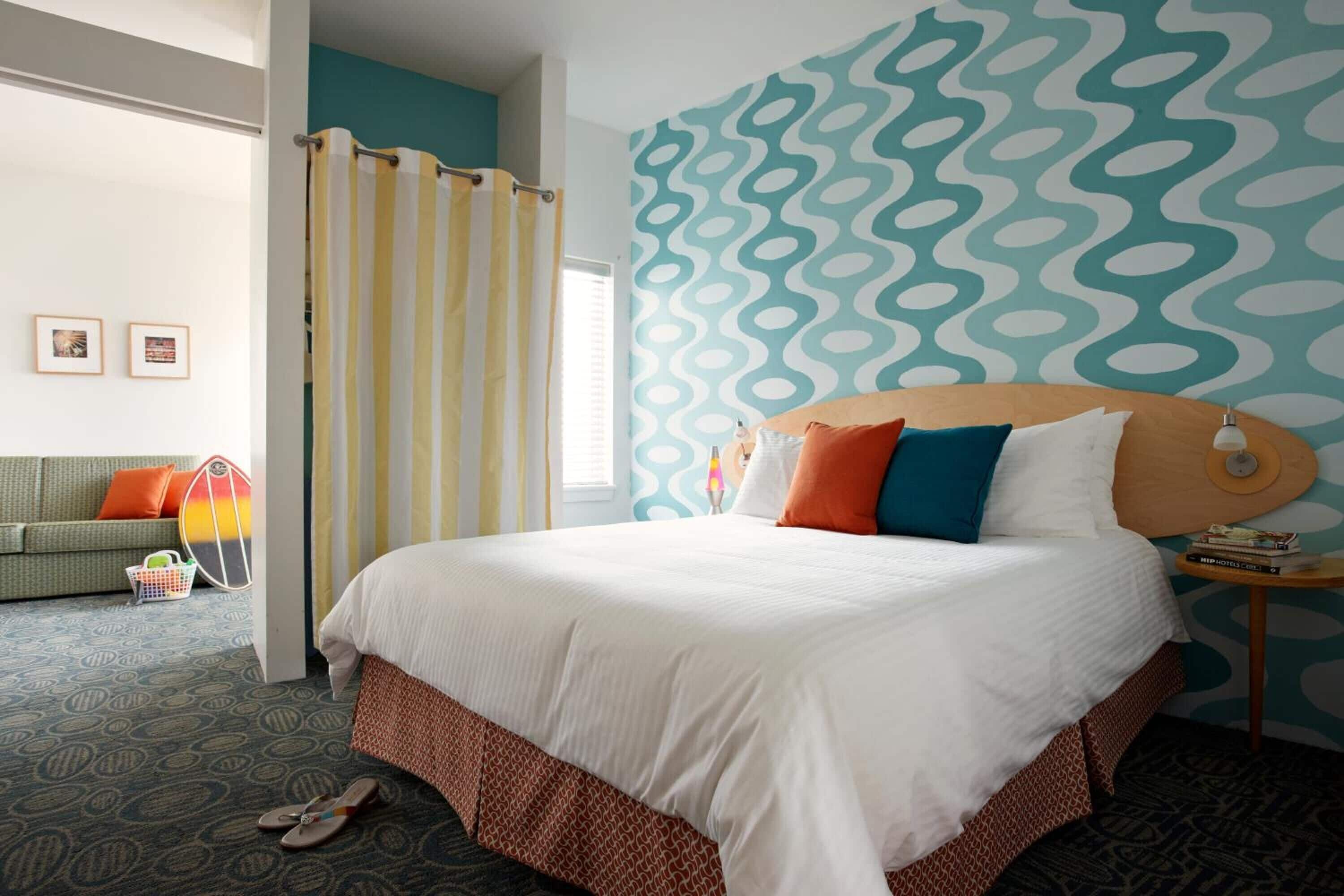 A hotel room with a large bed, a patterned wall, a sofa with cushions, and a small table with a lamp.
