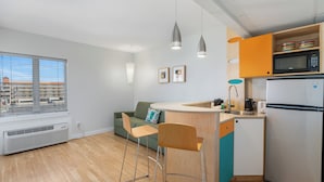 A modern kitchen with a built-in microwave, a refrigerator, and a window with blinds. There are two bar stools and a green sofa with a cushion. - Starlux Hotel (Wildwood)