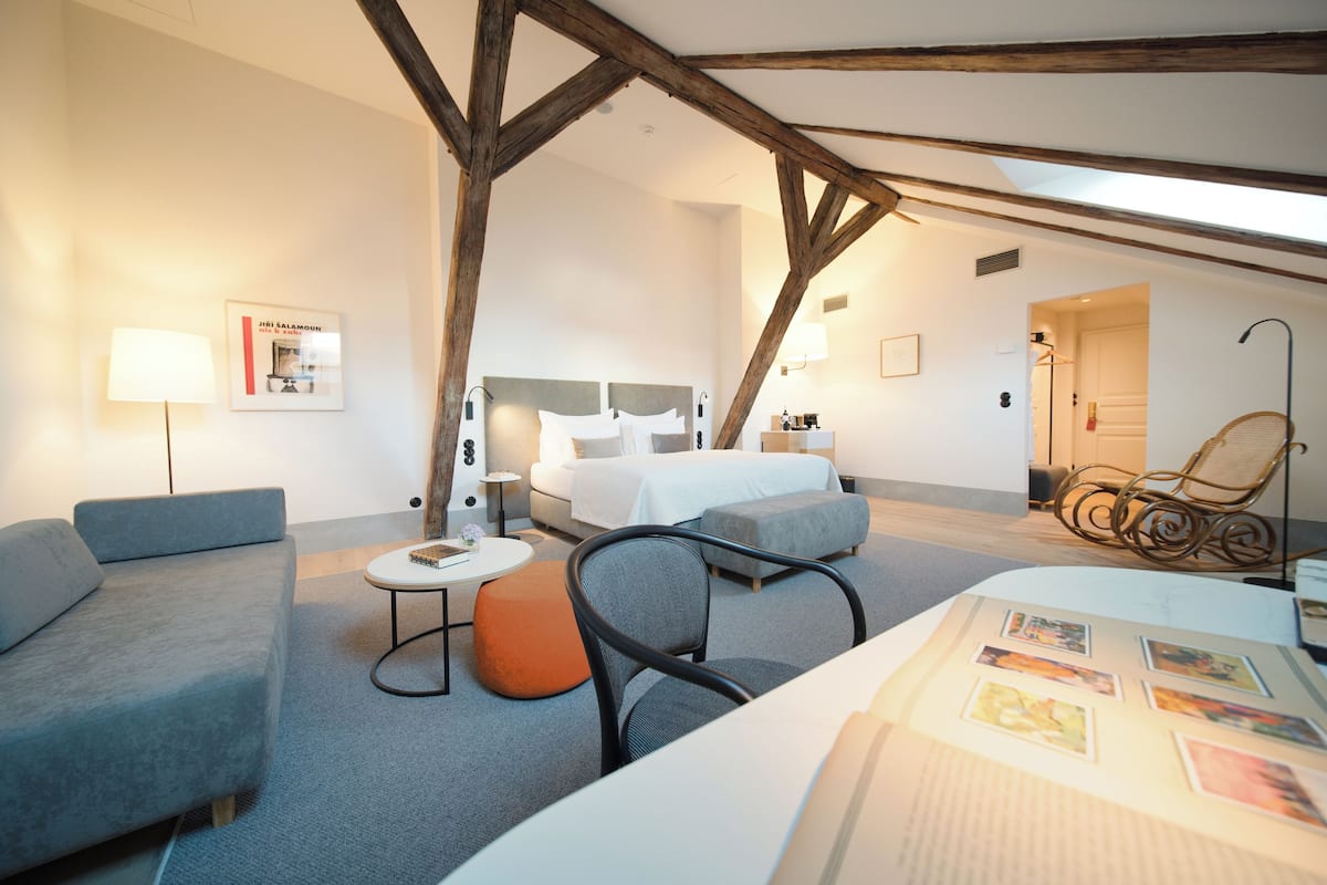 A modern hotel room with a loft-style ceiling, a bed with white linens, a grey sofa, a small round table, and a wicker chair.