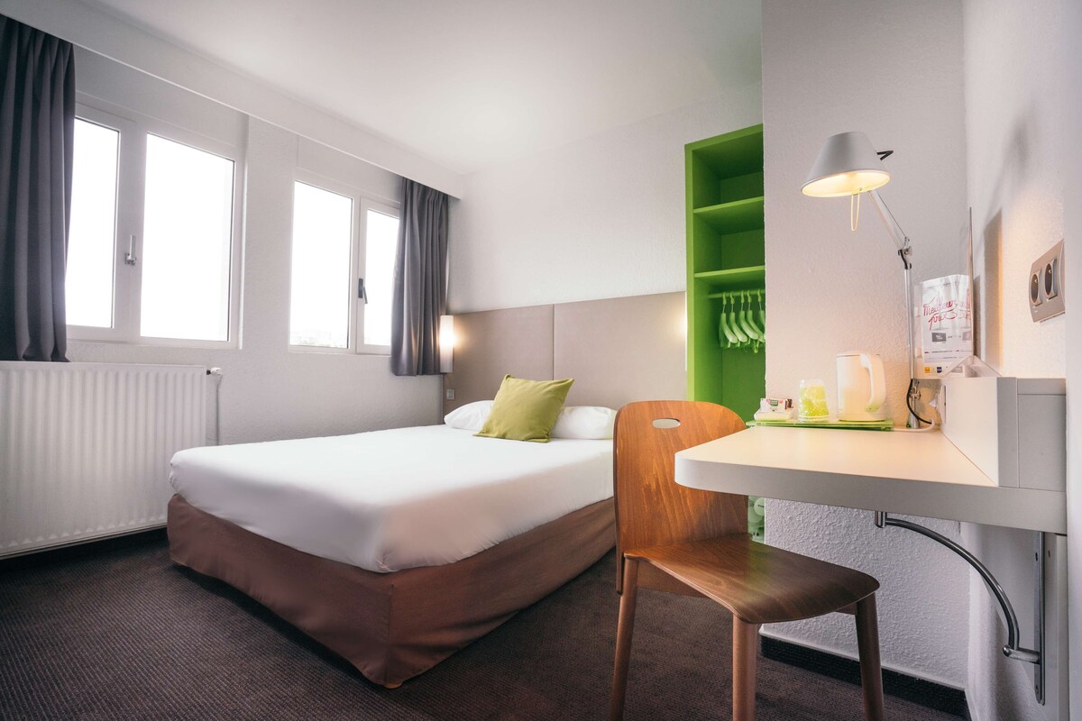 A hotel room with a bed, a desk, a chair, a green shelf, and a window with curtains.
