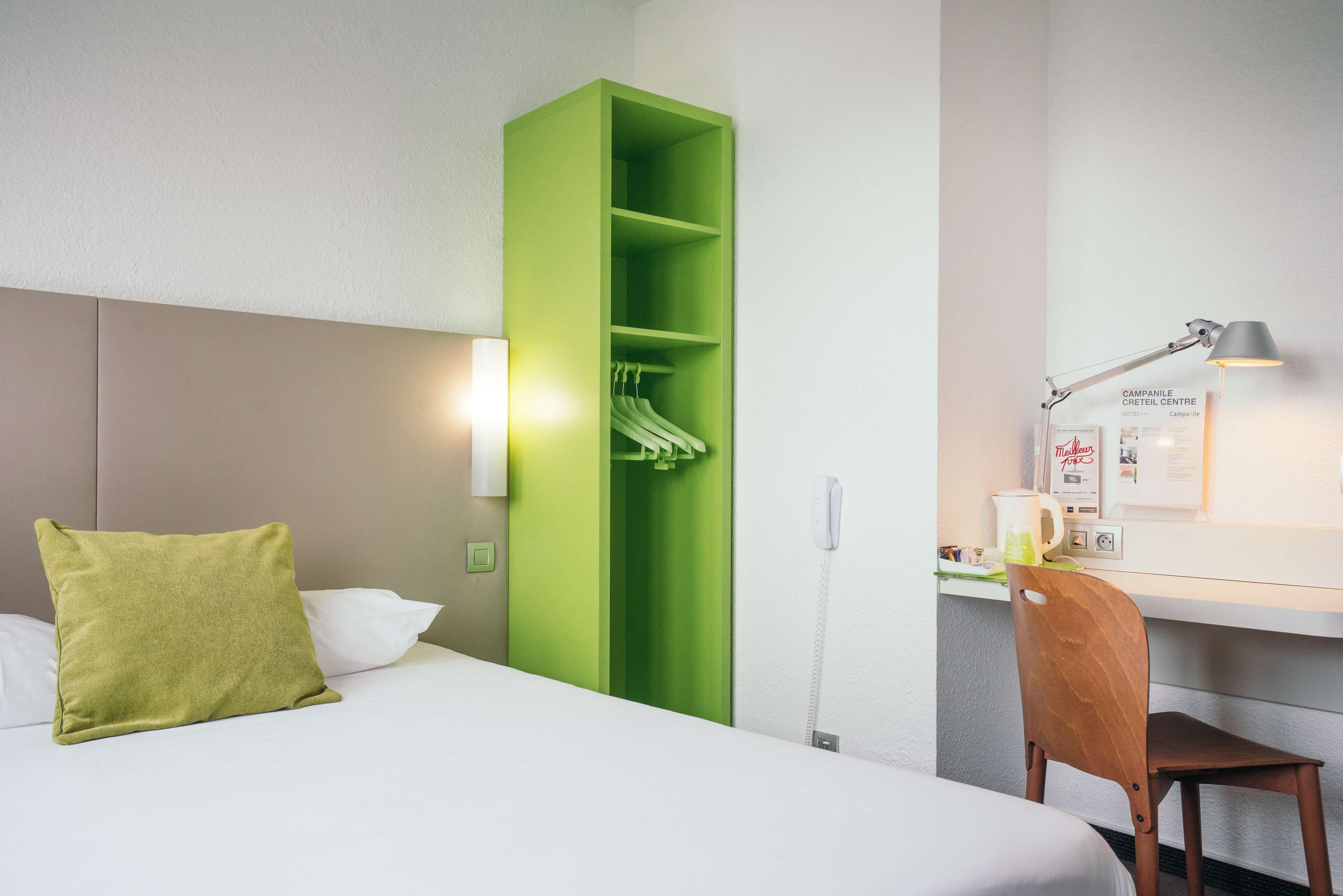 Una habitación de hotel con una cama, un armario verde, un escritorio con lámpara y una silla.