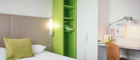 Una habitación de hotel con una cama, un armario verde, un escritorio con lámpara y una silla.