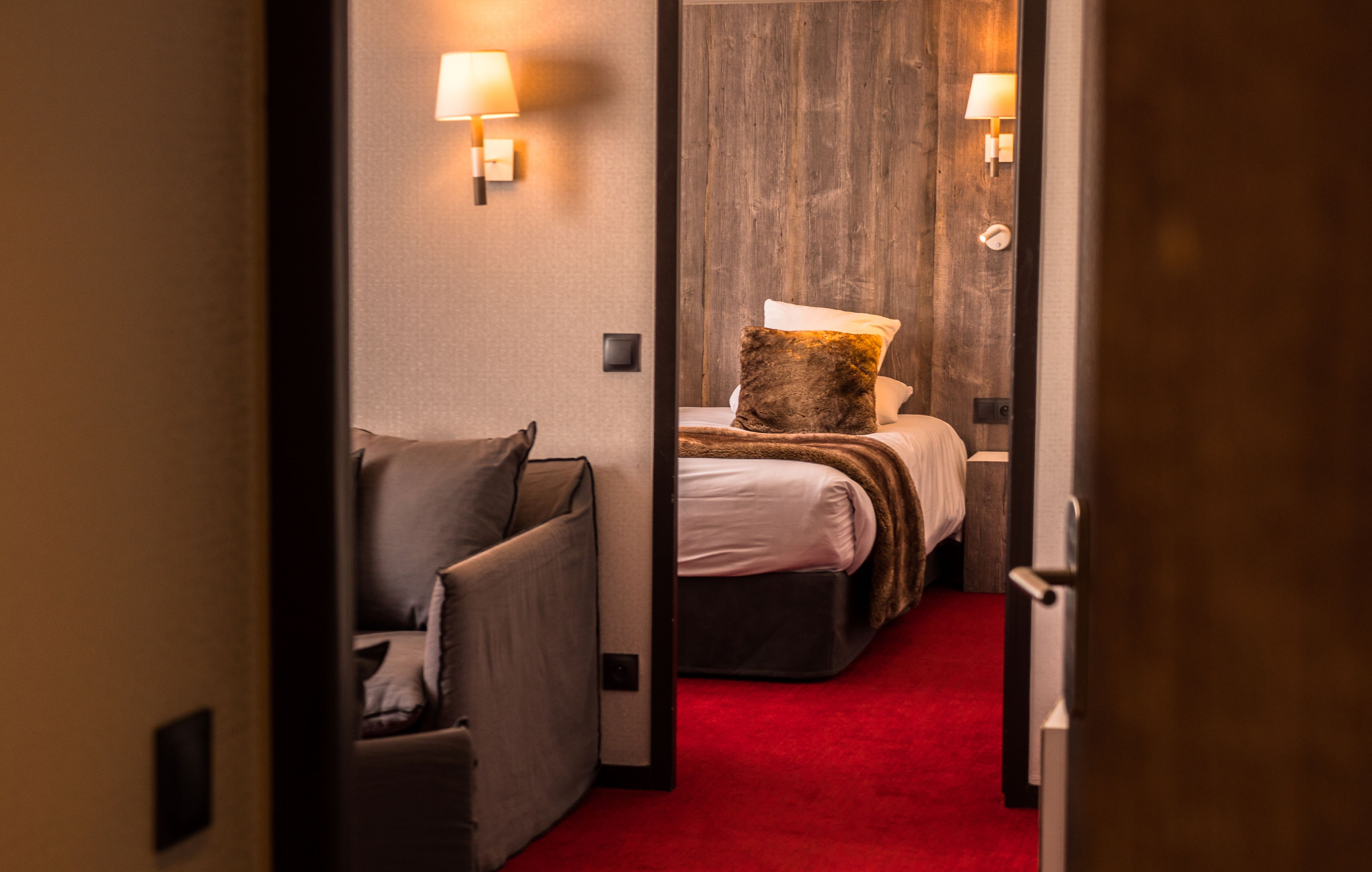 A hotel room with a red carpet, a bed with a brown pillow, a grey armchair, and two wall-mounted lamps.
