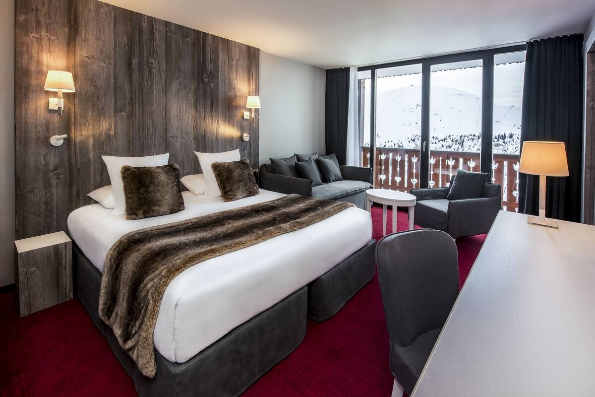 A hotel room with a large bed, a sofa, a desk, and a balcony with a view of snow-covered mountains.