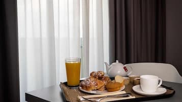 Ein Frühstückswagen mit Gebäck, Orangensaft, einem Teekessel und Tassen auf einem Tisch neben einem Fenster mit durchsichtigen Vorhängen.