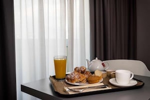 Bandeja de desayuno con pasteles, un vaso de jugo de naranja, una tetera y tazas sobre una mesa junto a una ventana con cortinas translúcidas.