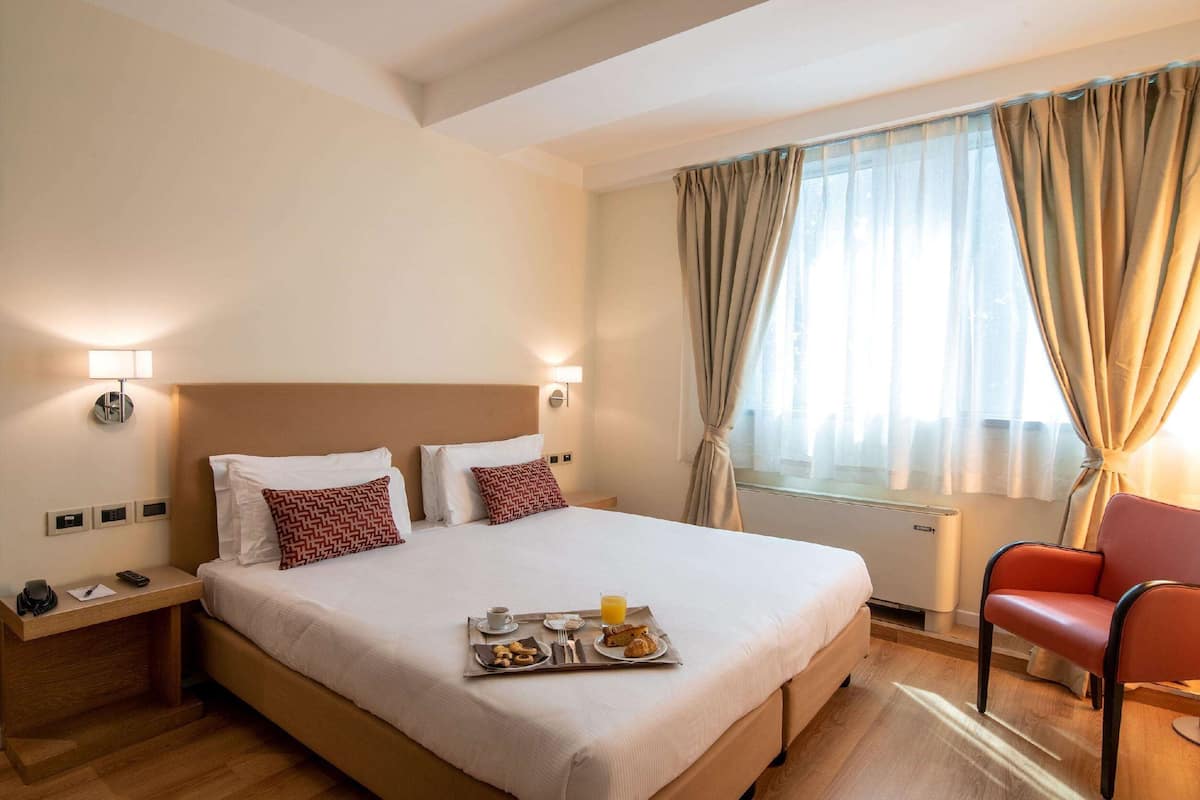 A hotel room with a large bed, a bedside table with a lamp, a chair, and a breakfast tray on the bed.