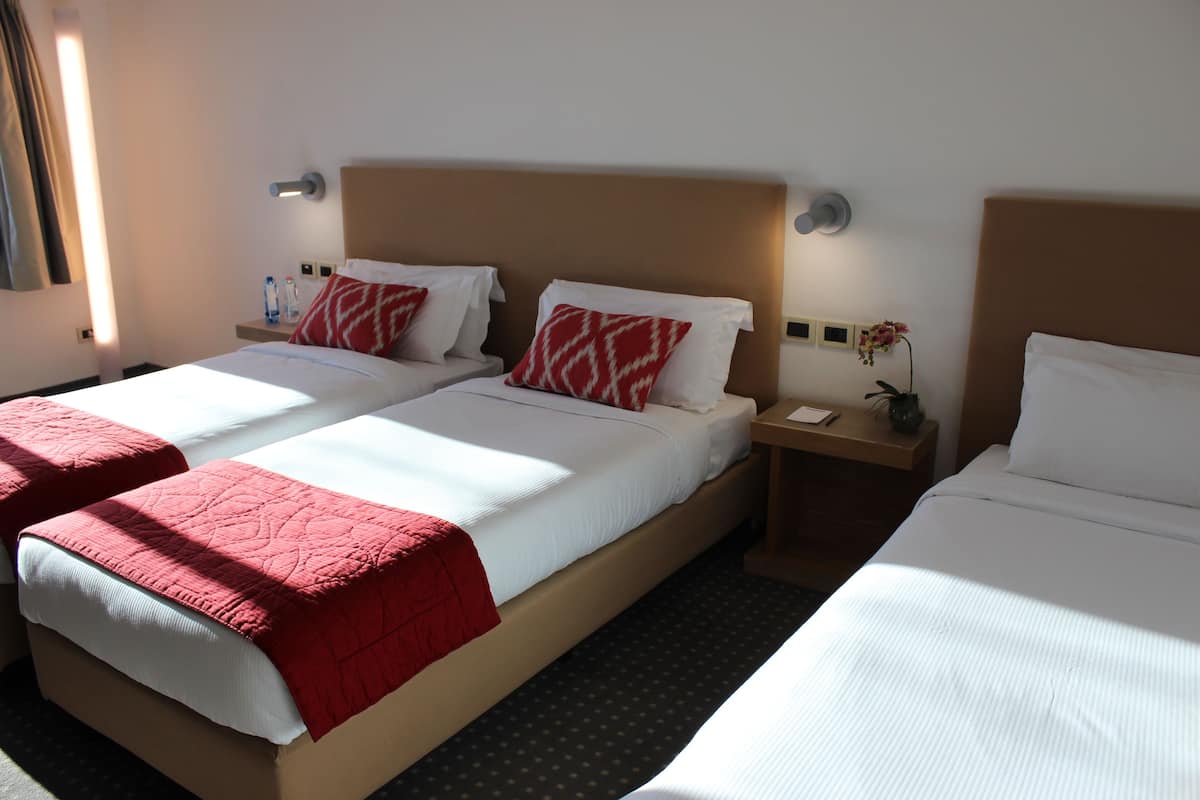 A hotel room with two single beds, each with a red and white patterned pillow, a wooden nightstand with a vase of flowers, and a wall-mounted light fixture.