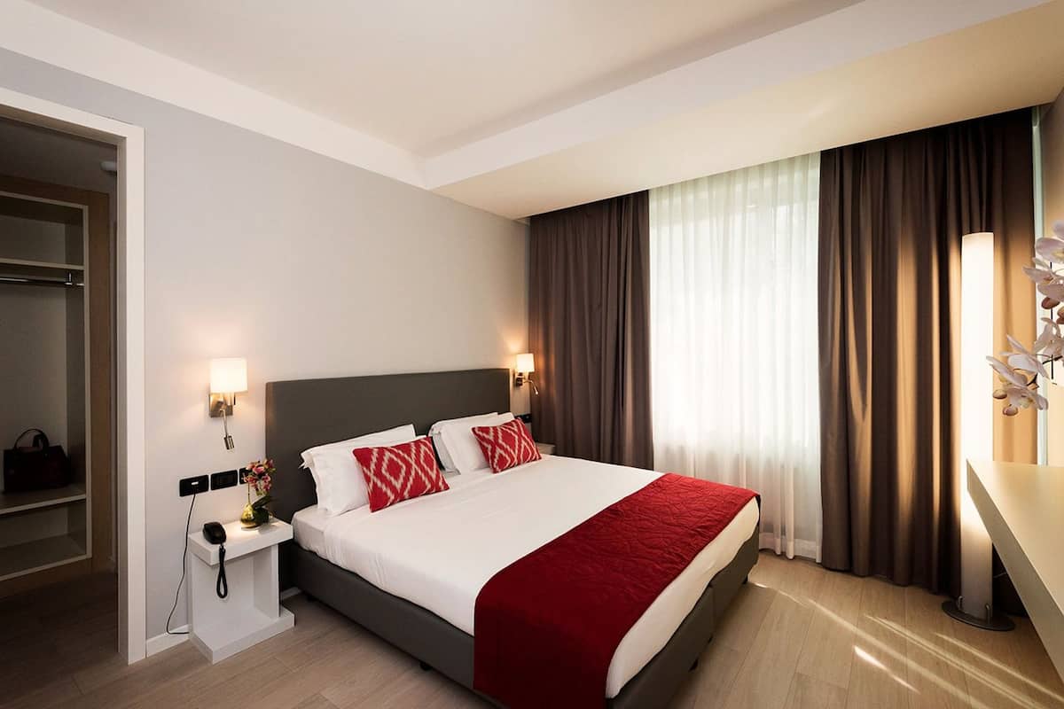 A hotel room with a bed, bedside lamps, a nightstand, a desk, and a window with curtains.