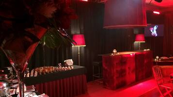 A dimly lit bar area with a red carpet, a bar counter, and a television screen in the background.