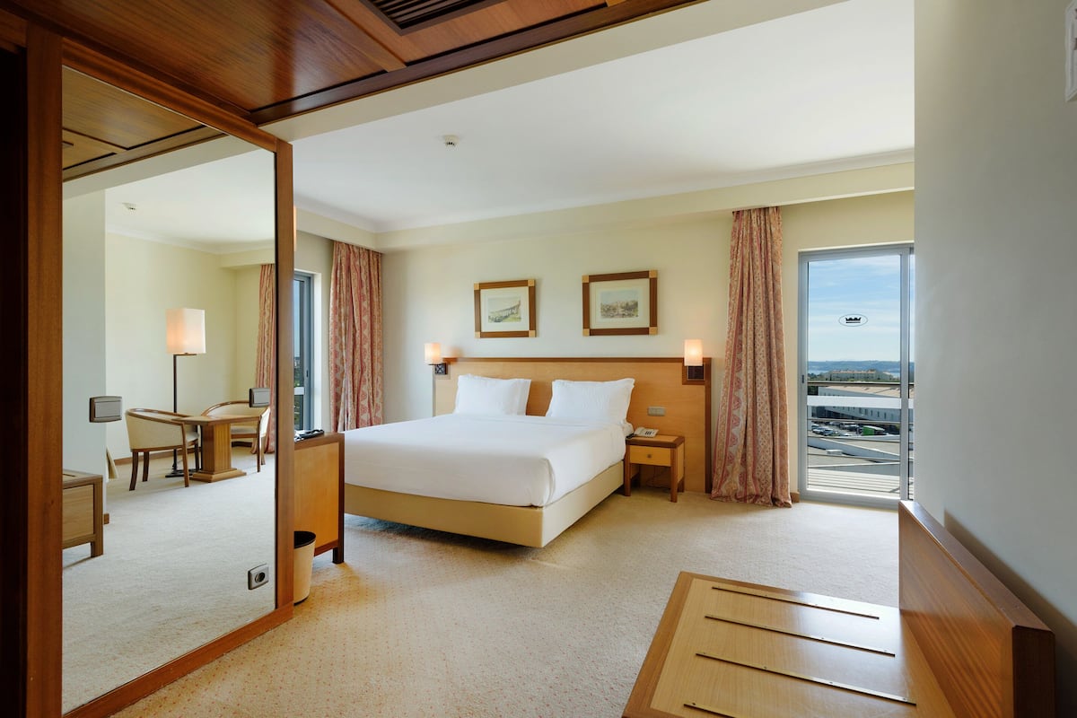 A hotel room with a large bed, a desk, a chair, a small table, a mirror, and a window with a view of the airport.