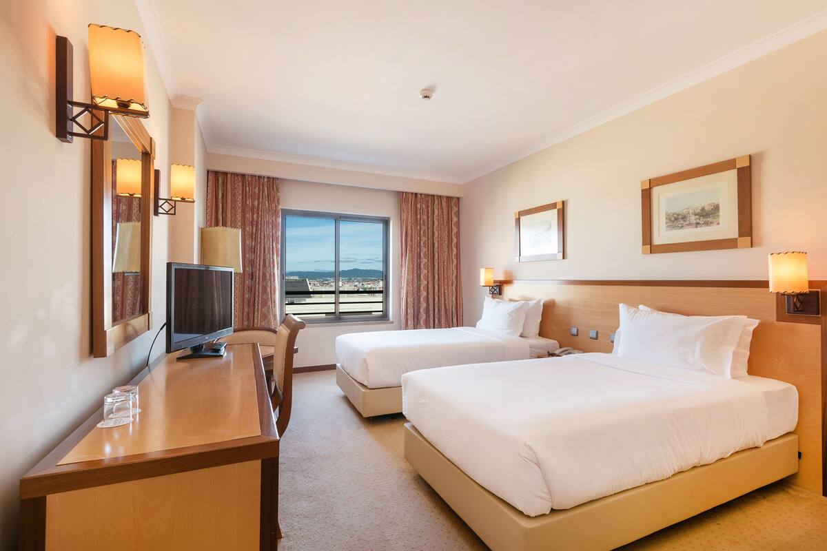 A hotel room with two beds, a desk, a TV, and a window with a view.