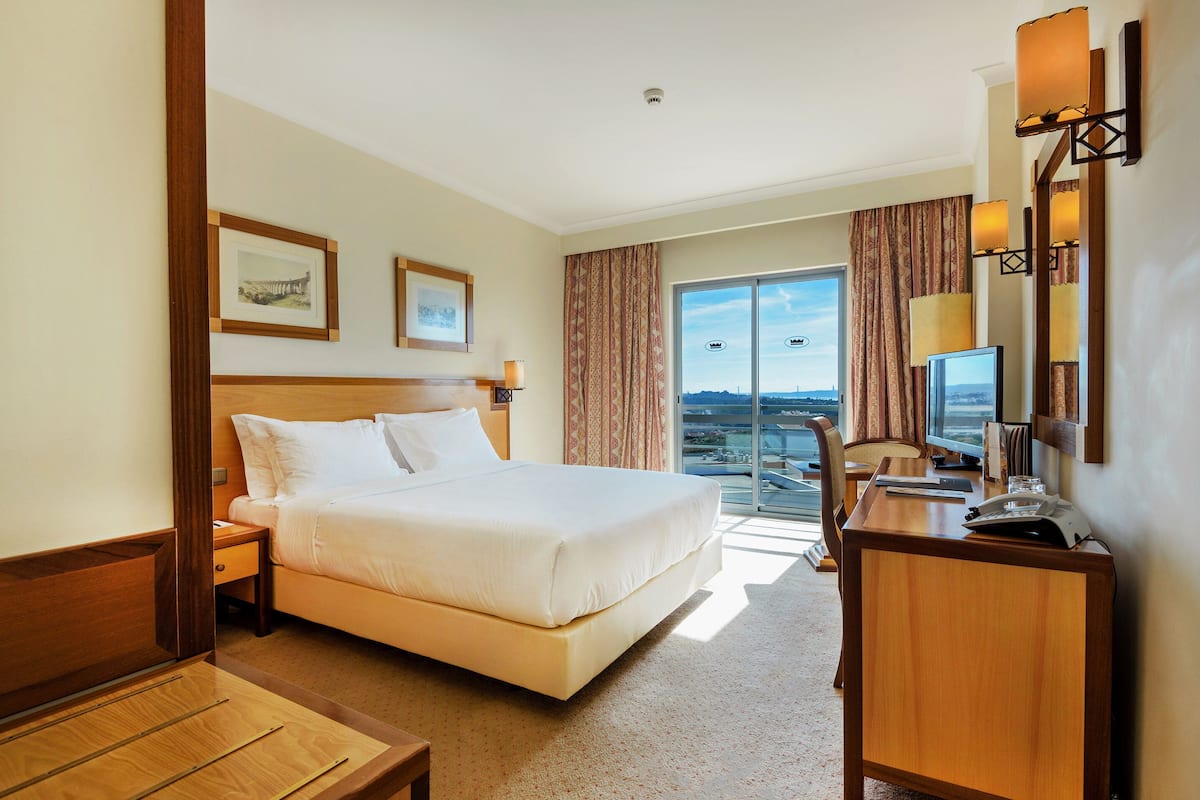 A hotel room with a large bed, a desk with a computer, a chair, and a view of the city through the window.