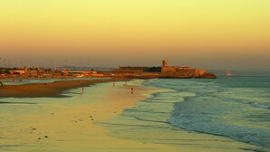 Escena costera con una playa arenosa, una estructura fortificada y un faro en la distancia.