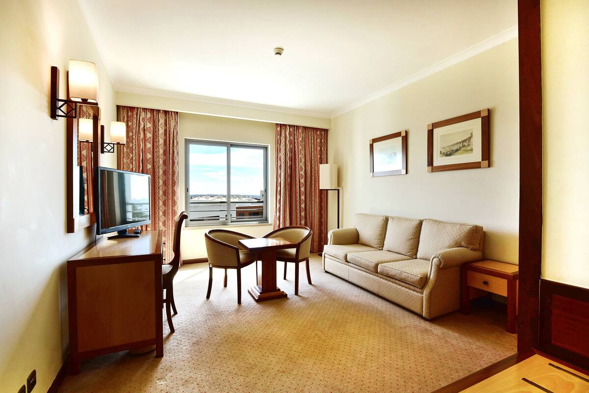 A hotel room with a TV, a dining table with chairs, a sofa, a small bedside table, and a window with curtains.