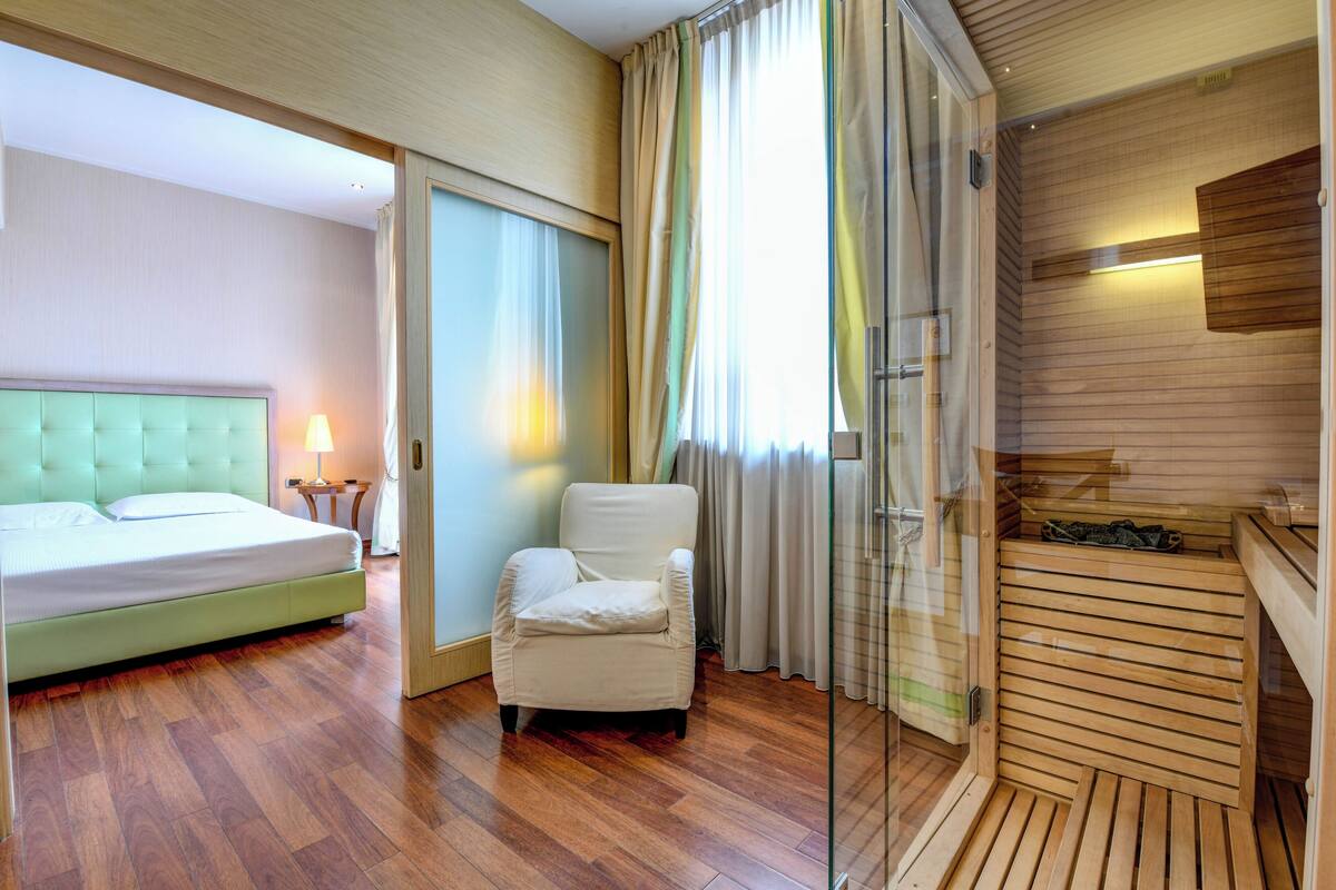 A hotel room with a bed, a chair, a glass door leading to a sauna, and wooden flooring.