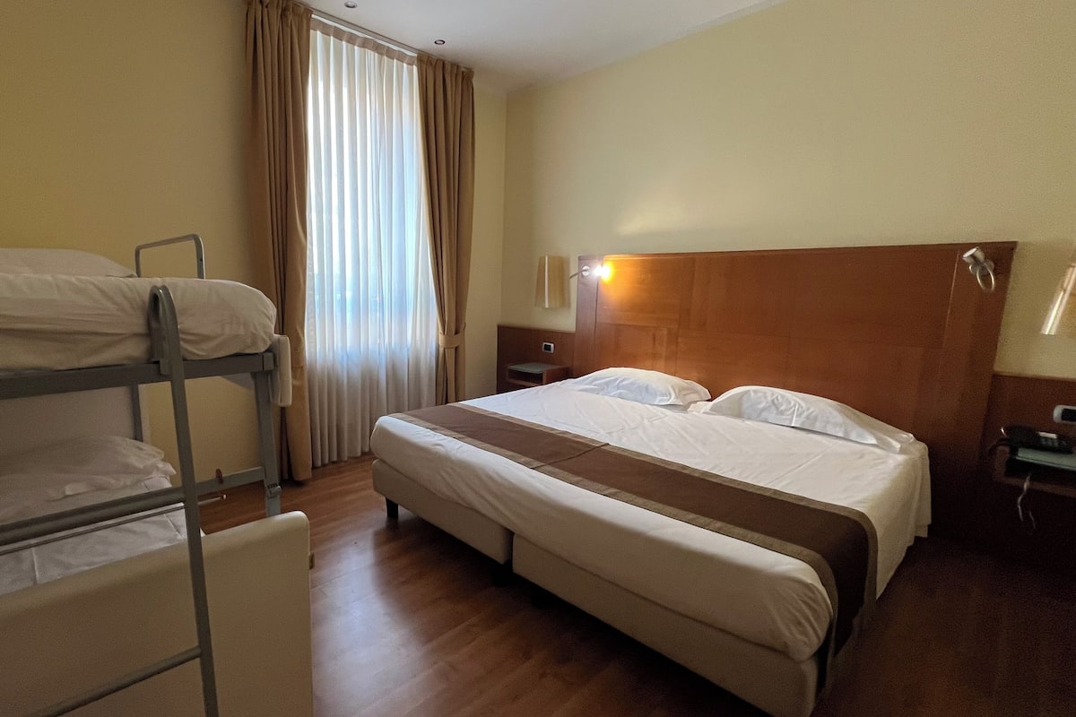 A hotel room with bunk beds, a single bed, a wooden headboard, a bedside table, and a window with curtains.