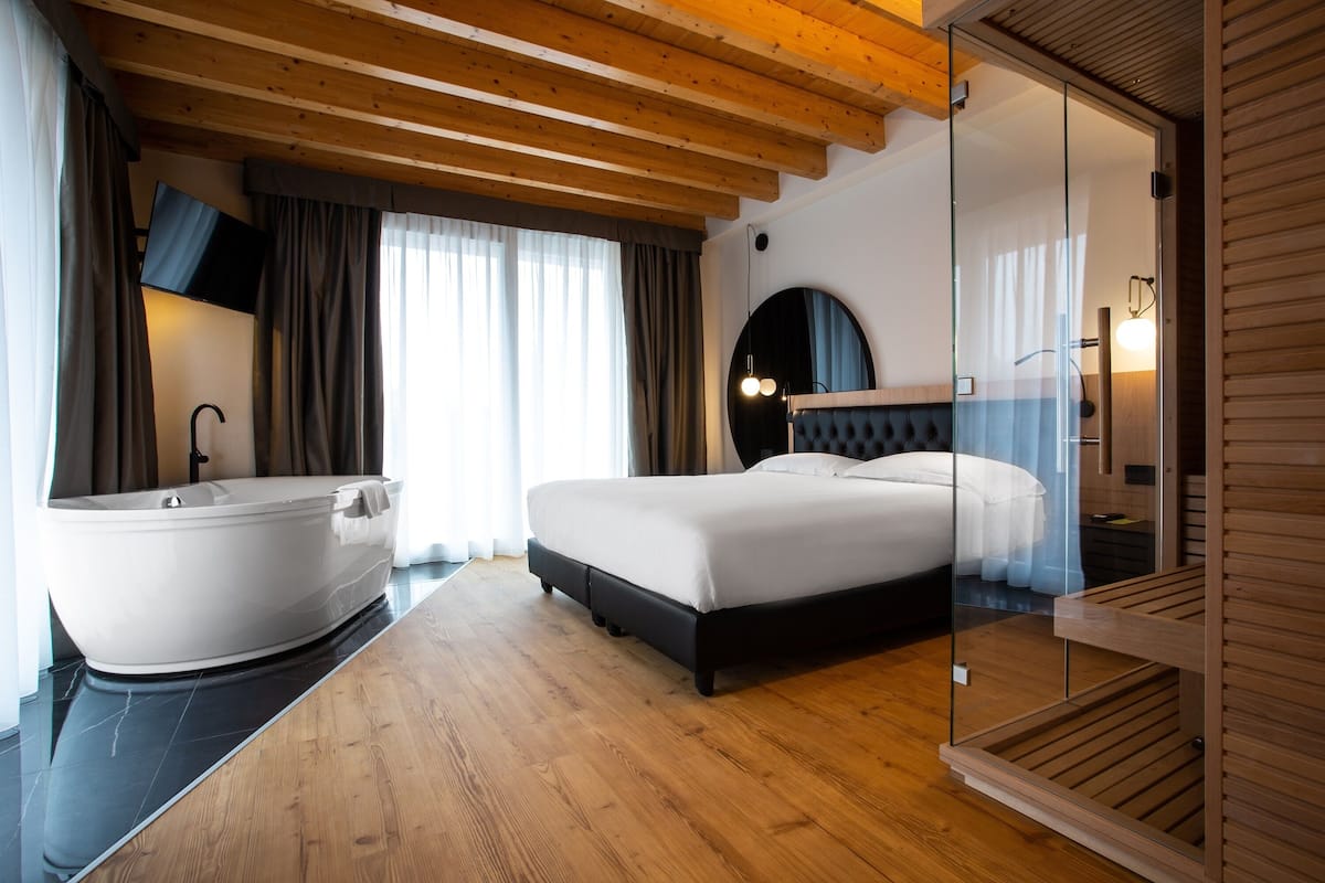 A modern hotel room with a large bathtub, a bed with a headboard, and a glass partition.