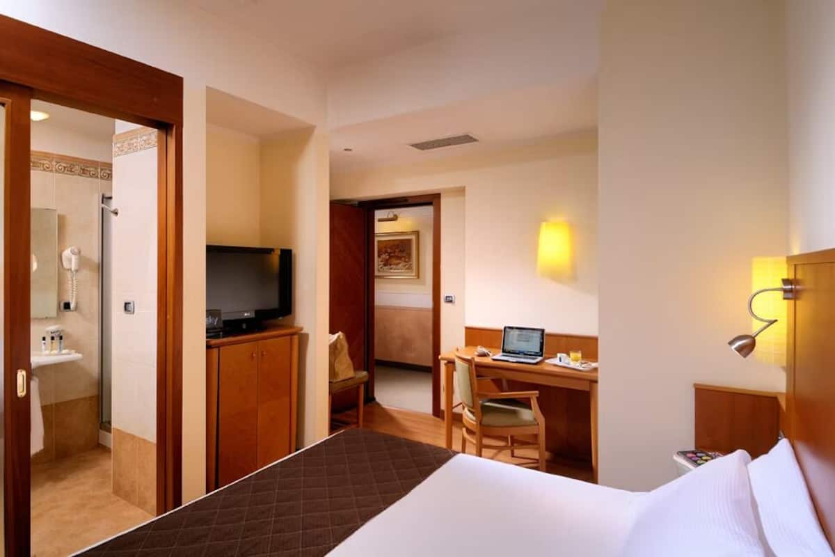 A hotel room with a bed, a desk with a laptop, a TV, and a shower area.