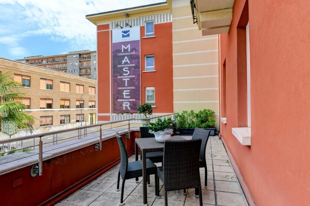 Un balcón con sillas de mimbre y una mesa, un edificio rojo con un letrero que dice 'MASTER', y vistas a otros edificios.