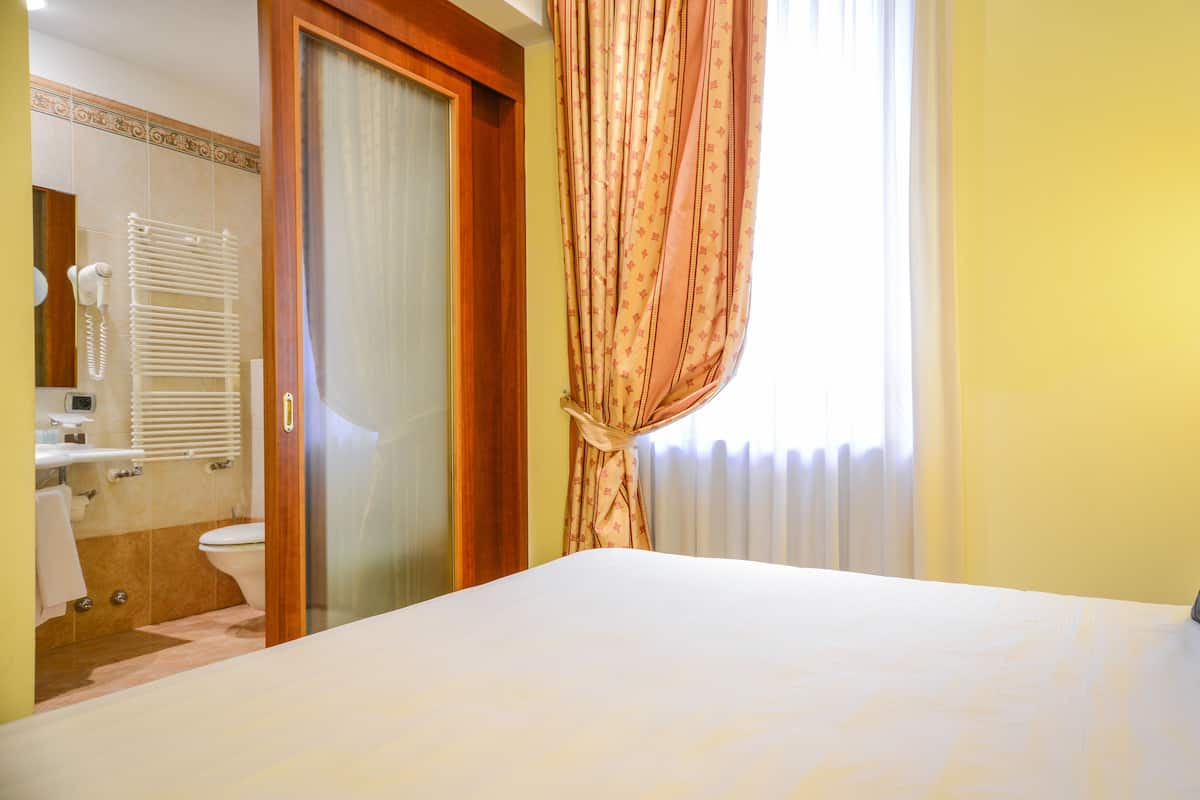 A hotel room with a bed, a bathroom with a toilet, a shower, and a window with curtains.