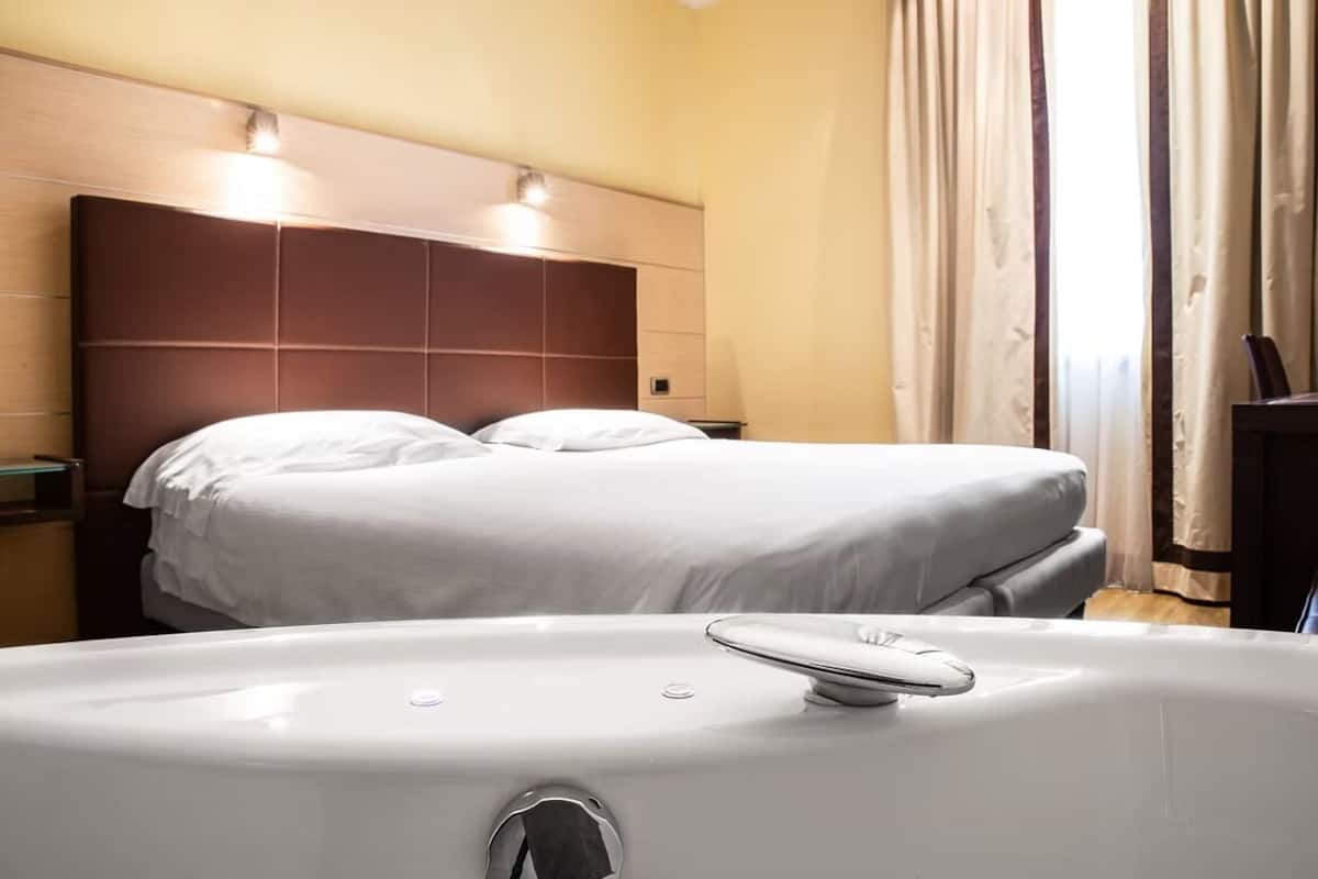 A hotel room with a large bed, a headboard with lights, a bathtub, and curtains.