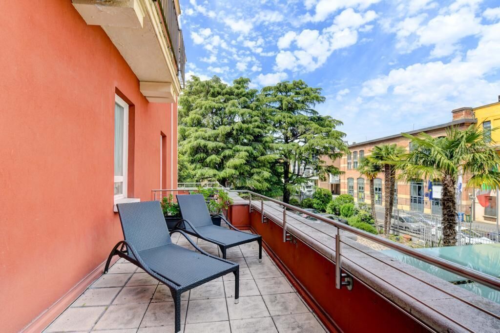 Un balcón con dos sillones de descanso, una pared de edificio rojo y vistas a árboles y edificios.