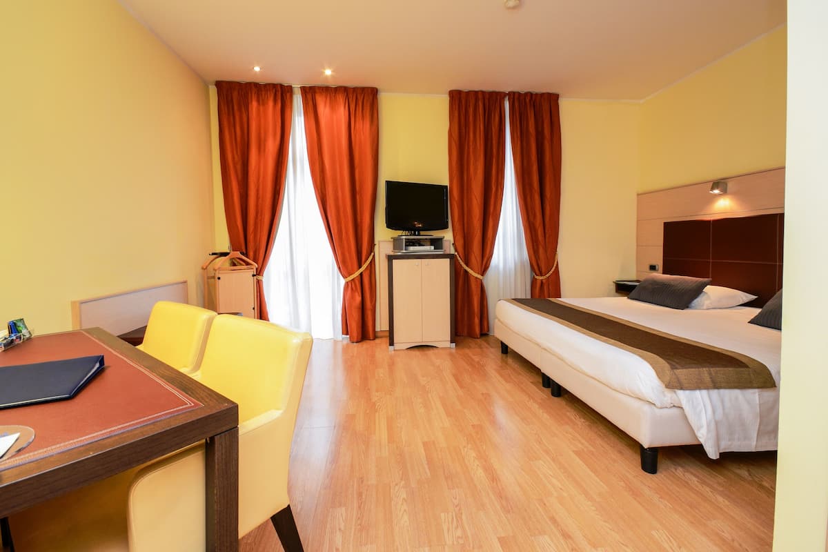 A hotel room with a bed, a desk with a chair, a television on a stand, and a wardrobe.