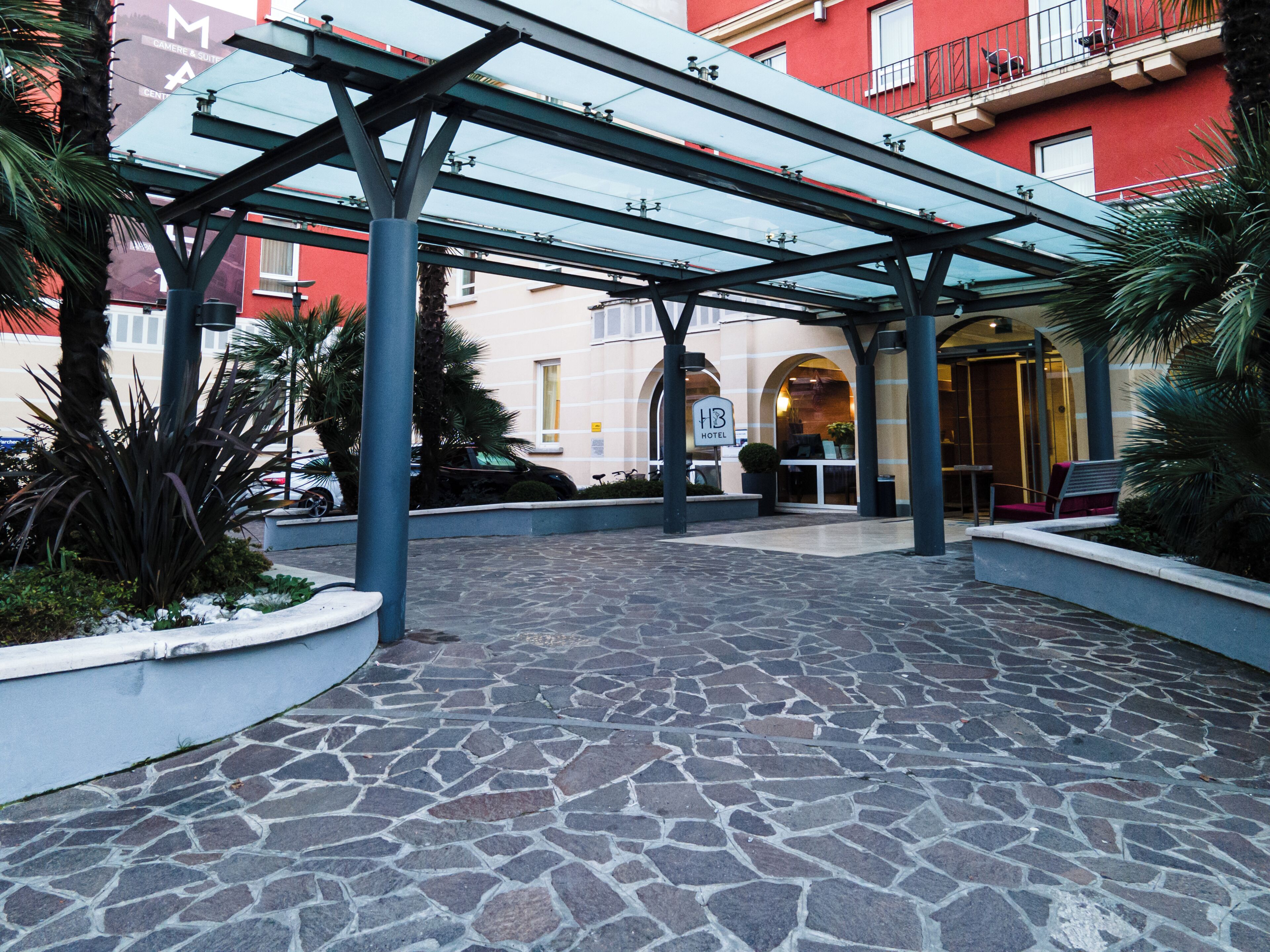 Entrada de hotel con un corredor cubierto, pavimento de piedra y un edificio rojo al fondo.