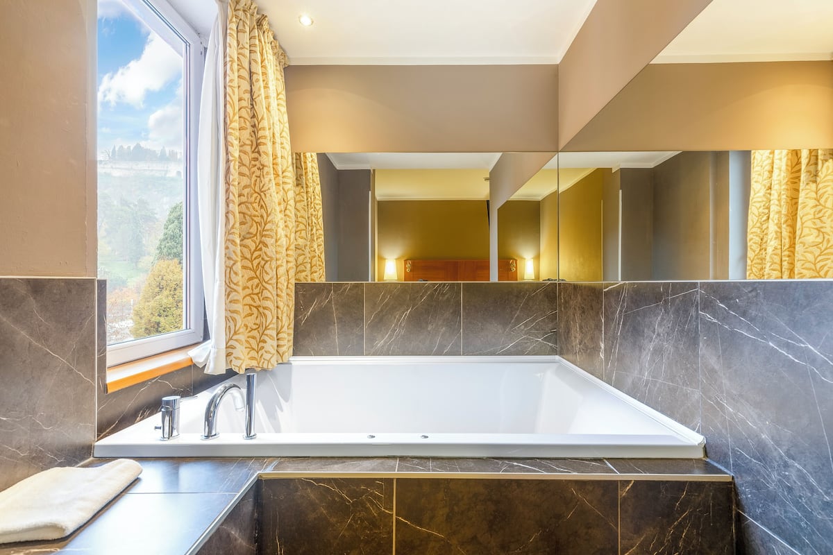 A modern bathroom with a large bathtub, a window with curtains, and a mirror reflecting the room.