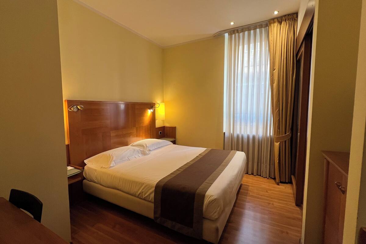 A hotel room with a wooden headboard, a bed with white linens, a bedside table, a lamp, and a window with curtains.
