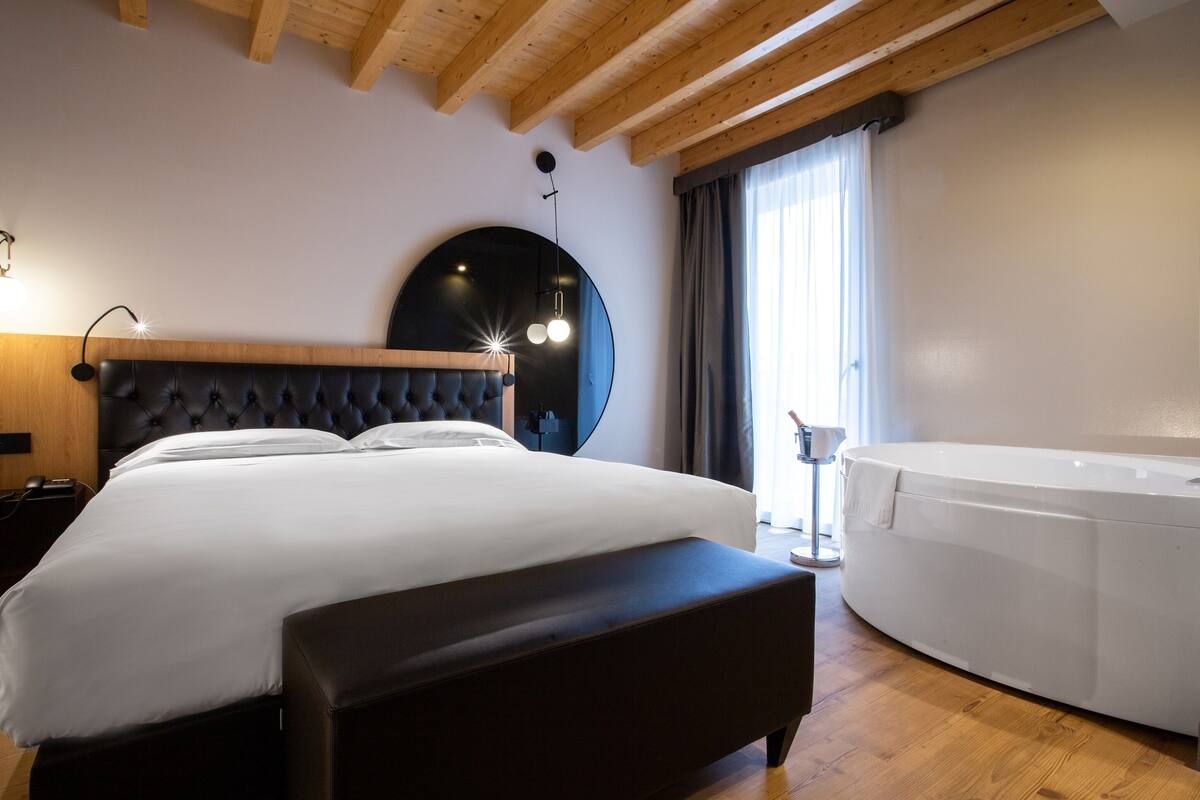 A modern hotel room with a large bed, a round mirror, a wooden ceiling, and a hot tub.