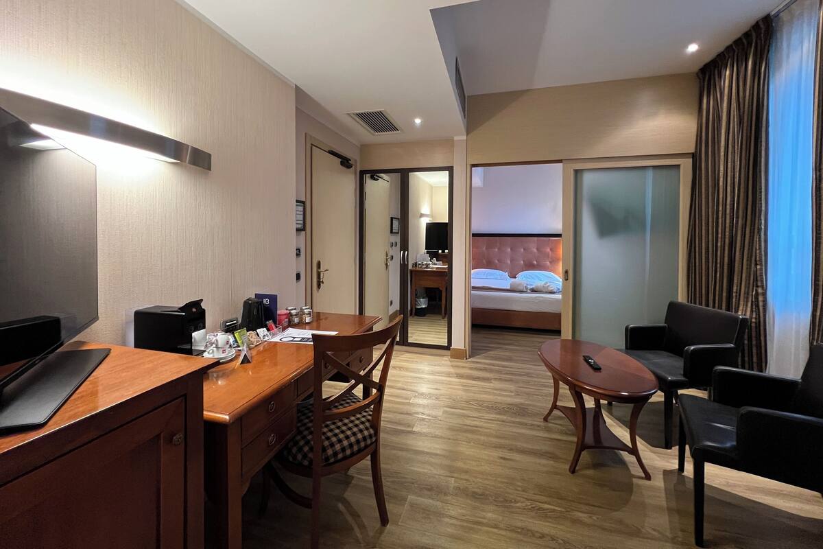 A hotel room with a wooden desk, a dining table, a bed with a headboard, and a view of the cityscape through the window.