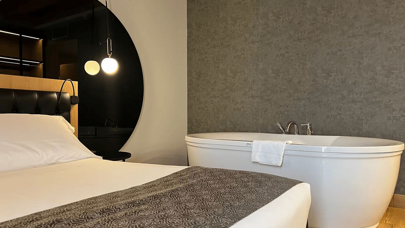A modern hotel room with a bed, a large round mirror, a round light fixture, and a freestanding bathtub.