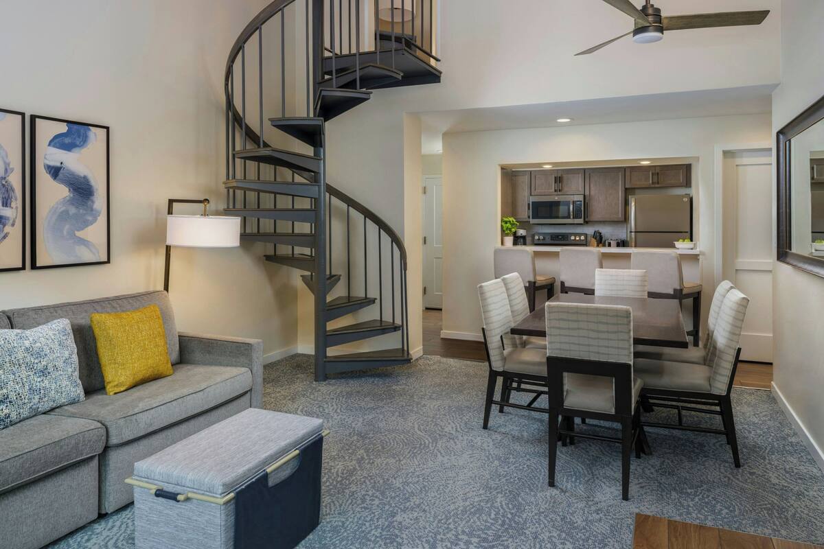 A modern living room with a gray sofa, a dining table with chairs, a spiral staircase, and a kitchen area in the background.