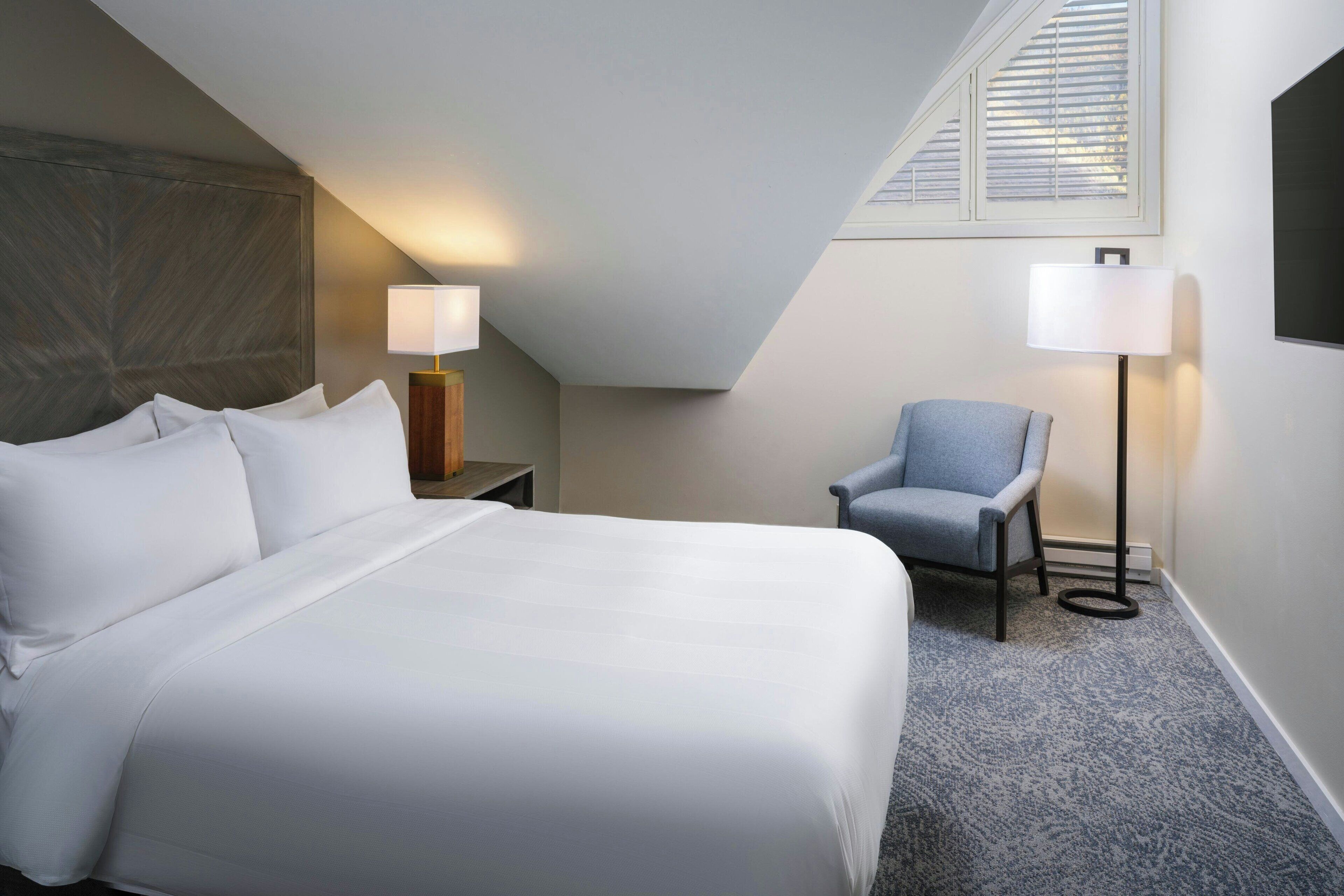 Une chambre d’hôtel avec un grand lit, deux lampes de chevet, une chaise et une fenêtre avec des stores.