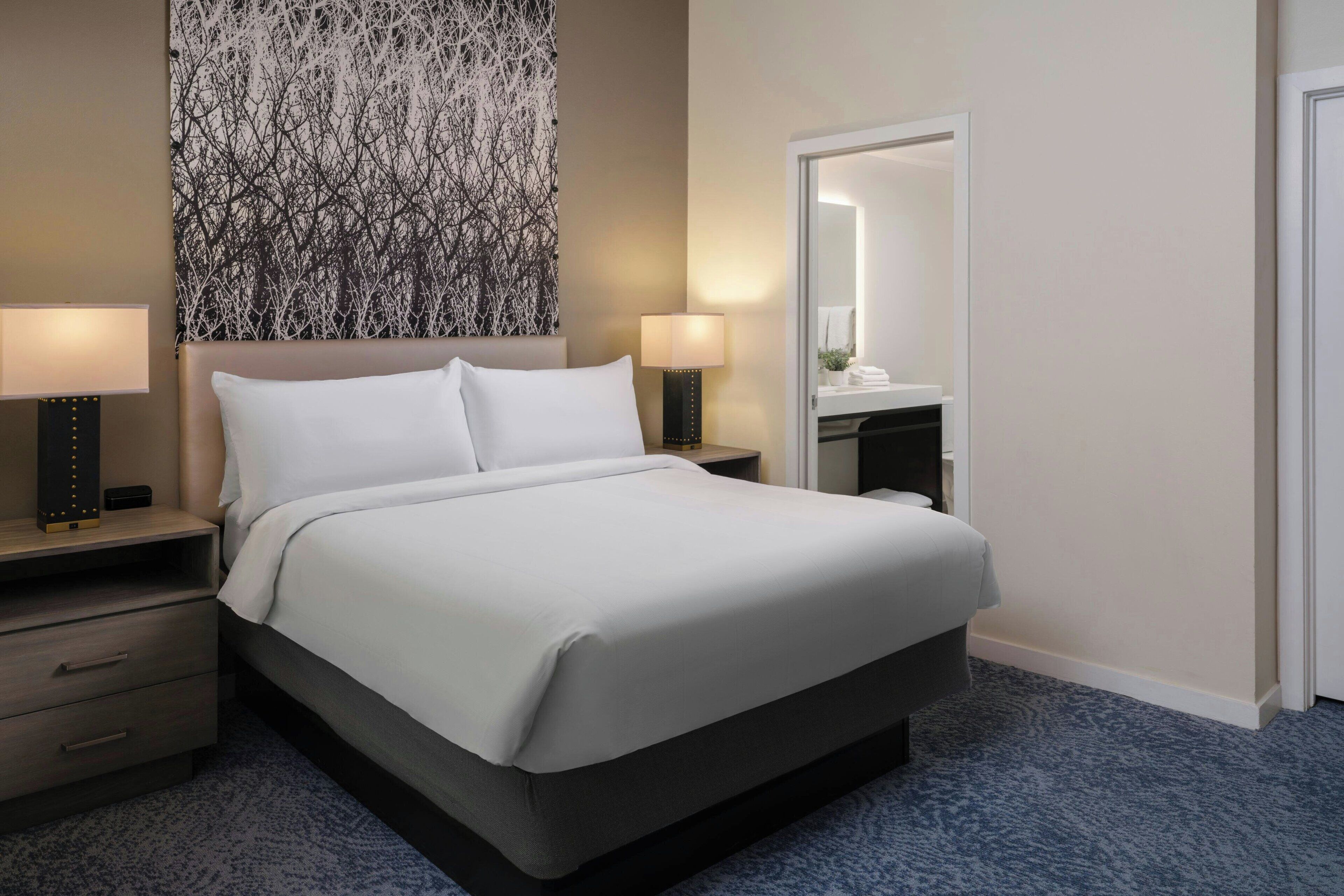 Una camera d'albergo con un letto grande, due comodini con lampade, uno specchio e un quadro appeso al muro.