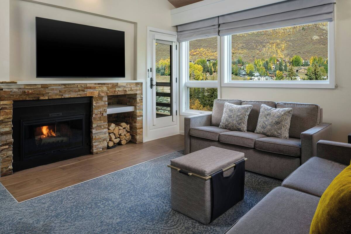 A living room with a fireplace, a sofa, a television, and a view of a mountainous landscape through large windows.