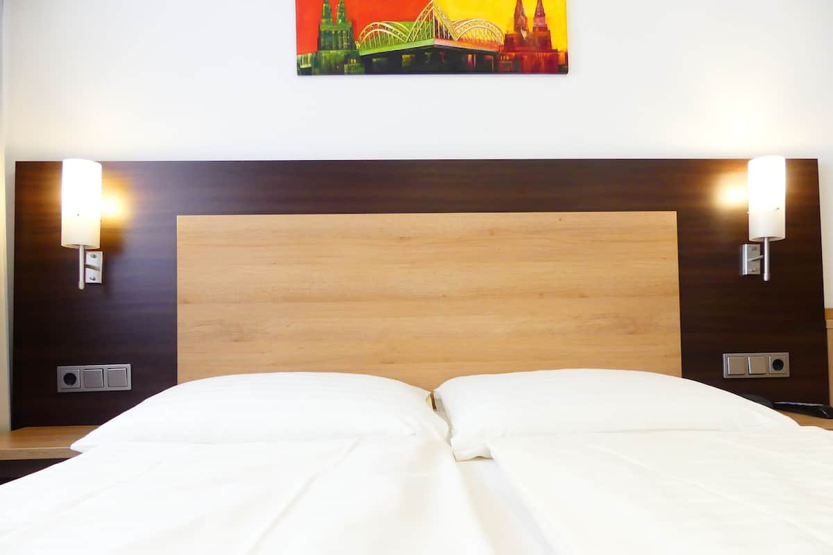 A hotel room with a wooden headboard, two wall-mounted light fixtures, and a cityscape painting above the bed.
