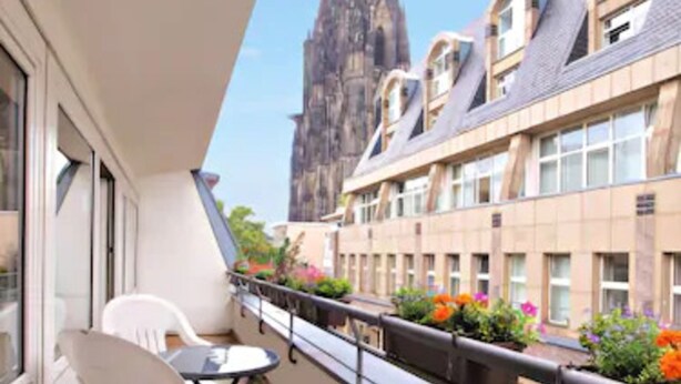 Ein Balkon mit Holzboden, weißen Stühlen und einem Tisch. Im Hintergrund ein Gebäude mit einem hohen Turm.